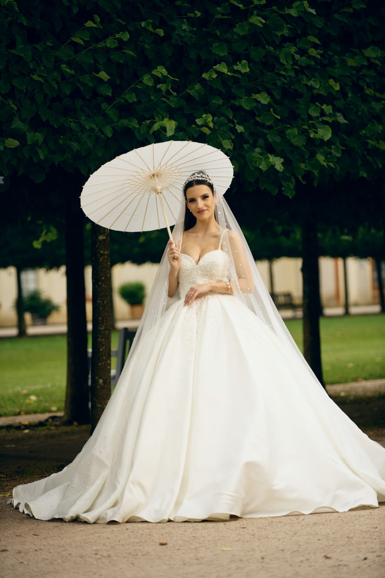 Braut in weißem Ballkleid mit Sonnenschirm in einer parkähnlichen Allee